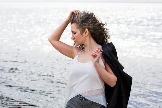 Portrait of a caucasian sad curly woman in a white blouse and black leather jacket standing on the seashore in the wind straightens her hair