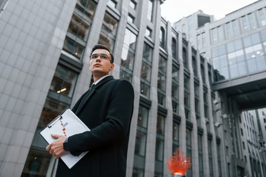 Grey building behind. Businessman in black suit and tie is outdoors in the city.