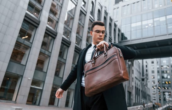 Grey building behind. Businessman in black suit and tie is outdoors in the city.