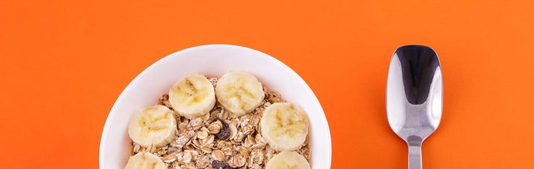 oatmeal in white plate with sliced banana and spoon on orange background, healthy food concept