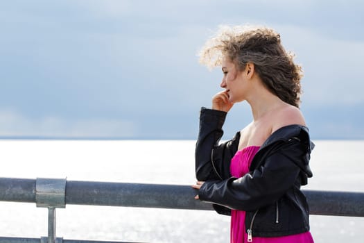 curly-haired Caucasian woman in a black leather jacket and a pink dress, stands on a bridge against the backdrop of a gloomy sky in the wind