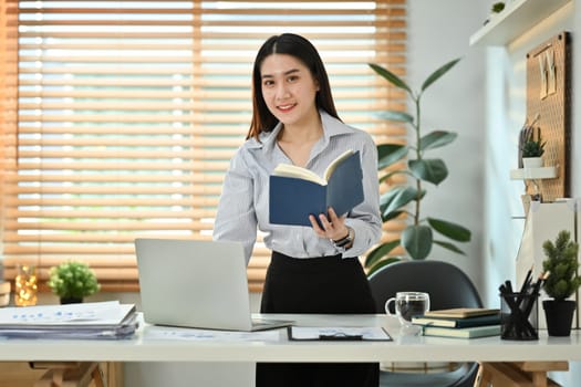 Beautiful young female entrepreneur holding diary planner standing at her working desk and using laptop computer.