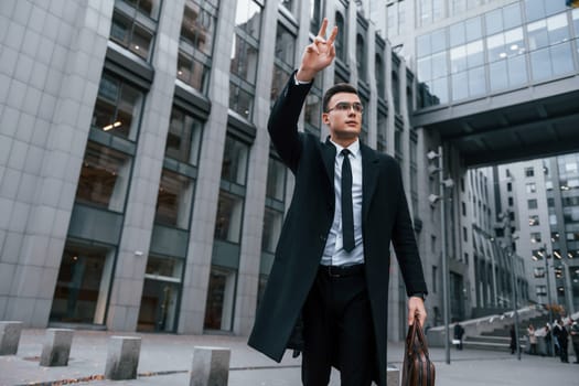 Raises his right hand. Businessman in black suit and tie is outdoors in the city.