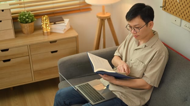 Image of millennial man sitting on couch in living room using laptop and checking her working schedule plan.