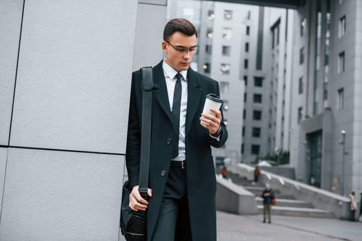 Standing near the building. Businessman in black suit and tie is outdoors in the city.