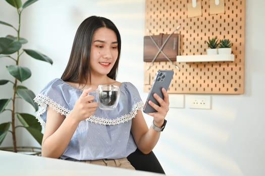 Happy millennial woman drinking coffee and reading massage on mobile phone. Technology, communicating concept.