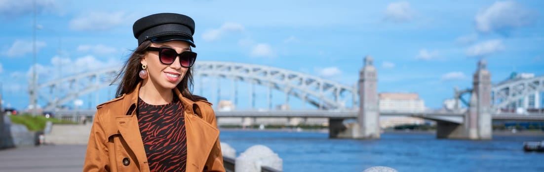 Happy young woman of Caucasian ethnicity in a light brown coat and black cap posing on the embankment against the backdrop of the bridge and the blue sky.