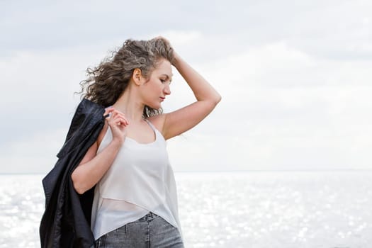 Portrait of a caucasian sad curly woman in a white blouse and black leather jacket standing on the seashore in the wind straightens her hair