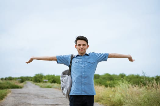 Happy young man posing with raised arms, standing on the road and looking into the distance. Happy guy in a blue shirt with a backpack. Free student enjoys vacation