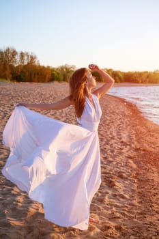 A beautiful young woman of Caucasian appearance with dyed hair posing on the seashore in the sun at sunset in a white dress in the wind. Romantic slender girl in nature enjoys relaxation