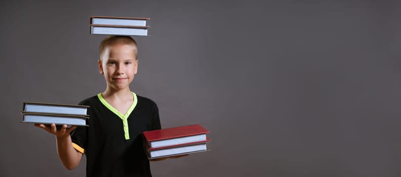 little blond boy in a black T-shirt hold stacks of books in your hands and on your head. the child is looking at the camera, isolated on a gray background.