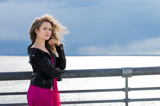 curly-haired Caucasian woman in a black leather jacket and a pink dress, stands on a bridge against the backdrop of a gloomy sky in the wind