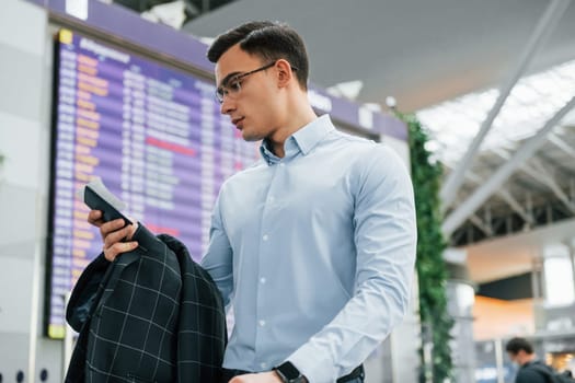 Holding phone. Young businessman in formal clothes is in the airport at daytime.