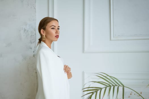 portrait of a beautiful young blonde woman with large gold earrings and a white jacket on the background of the white interior of the living room. The model is taken in daylight and looks away.