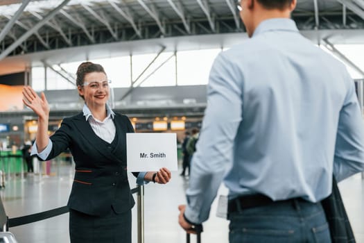 Meet by the woman with text. Young businessman in formal clothes is in the airport at daytime.