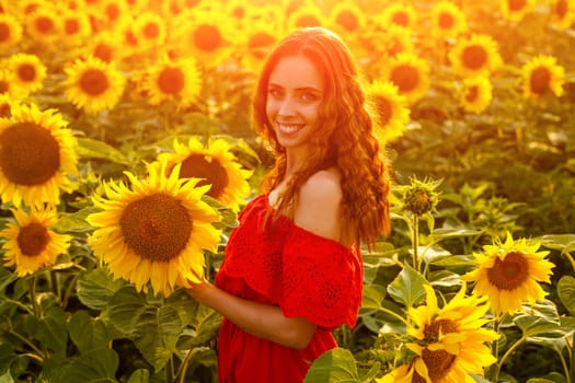 Female portrait of young woman in field of blooming sunflowers in rays of sunset . Cute girl of Caucasian appearance in evening, free and happy in red dress, stands in a field of bright sunflowers