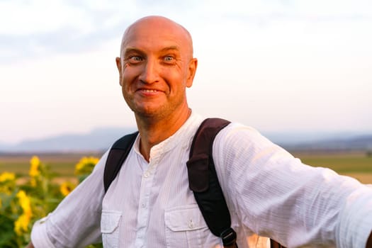 Close-up of a cheerful young man in a white t-shirt with a backpack stands and smiles in a field of sunflowers. Cute caucasian bald man outdoors during sunset