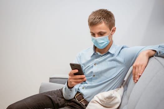 young man of European appearance in casual clothes, at home on the couch in a medical mask communicates by video communication on the phone, the concept of remote online communication on the Internet