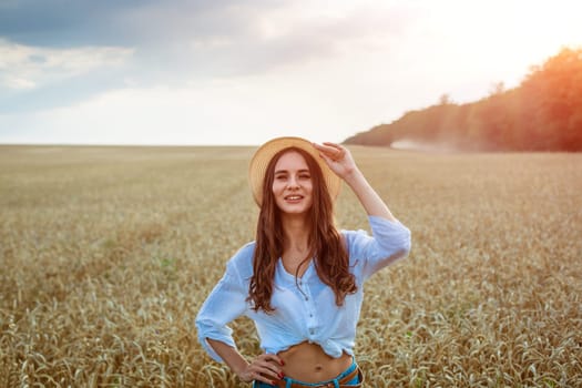 Happy girl sits in wheat field in straw hat. Cute young woman of Caucasian ethnicity in casual clothes enjoys ripe golden wheat in a field. Free woman concept