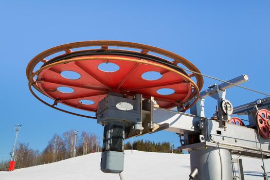 Large spinning wheel with electric cable hoist motor in a ski resort. The drive mechanism against the blue sky. Mountain slope equipment for snowboarding. Ski tours on a sunny day.