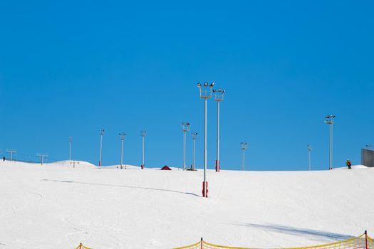 Ski resort, snow slope, trail with towers and floodlights. Mountain slope for skiing and snowboarding. Against the background of the blue sky. The summit of the rise and ski tours on a sunny day.