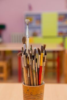 Artistic brushes stand in a wooden glass close-up. The objects for drawing with paints are collected together. In the background there is a table and cabinet out of focus. Warm soft daylight.