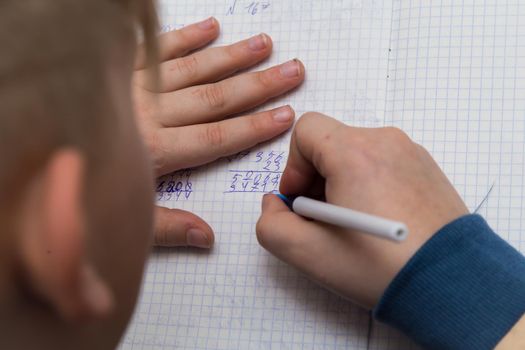 The boys hands with a ballpoint pen solve a close-up math example. The student completes the task in teradi. A schoolboy teaches lessons at a school desk. Warm soft daylight.