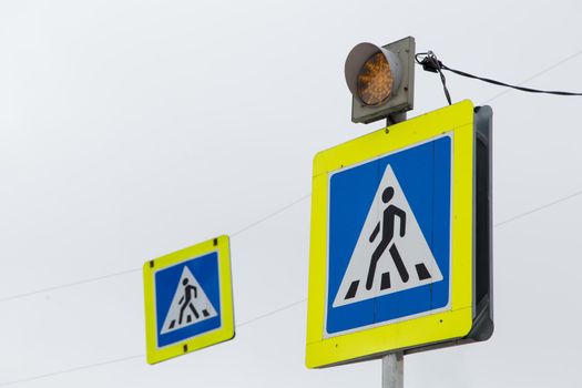 Road sign white triangle with a man in a blue square means a pedestrian crossing. The yellow traffic light warning light is on. Close-up on the background of the gray sky. There is a copy space.