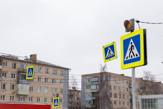 Road signs man in a blue square, means a pedestrian crossing. The yellow traffic light warning light is on. Close-up on the background of residential buildings. There is a copy space.