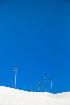 Ski resort, gentle snow slope with lighting towers and a moon in the sky. Mountain slope for skiing and snowboarding. Against the background of the blue sky. Ski tours on a sunny day.