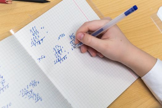 The girls hand with a fountain pen with blue ink solves examples in mathematics. The student completes the task in teradi. A schoolboy teaches lessons at a school desk. Warm soft daylight.