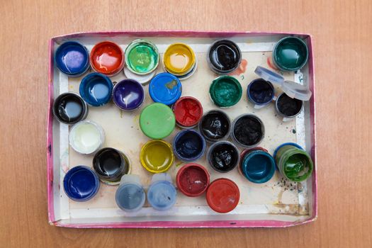 A box with old open jars of gouache for painting. Top view of half-empty jars of gouache paints. Flat layout on the table.