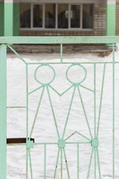 An iron lattice with metal figures of people of light green color. A gate made of metal rods with a latch against the background of the school building out of focus. Warm winter day, there is snow.