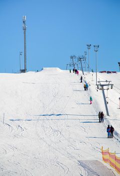 Cable car with people going to the top at the ski resort. Lighting poles and cell tower against the blue sky. Mountain slope lighting for snowboarding. Ski tours on a sunny day.