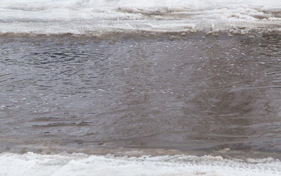 A deep puddle of ice and snow along the edges on the road in the city. Abnormal weather concept. There is copy space. The New Year and Christmas celebrations are over. Remains of last year's snow.