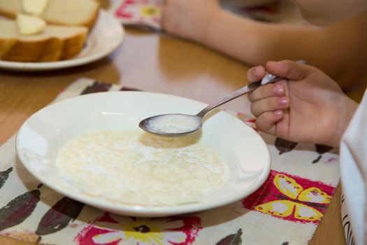 Close-up of a boy's hand with a spoon of porridge over a plate. A boy is having breakfast at the dinner table. A plate of porridge is on a napkin.