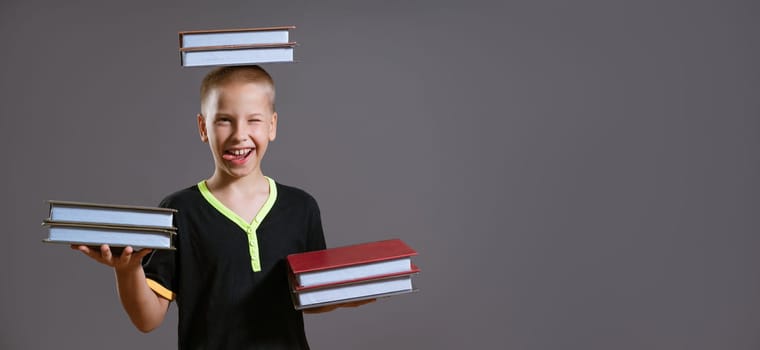 Funny Caucasian boy holding books in his hands and on his head. Shows her tongue on a gray background in casual wear. Back to school concept