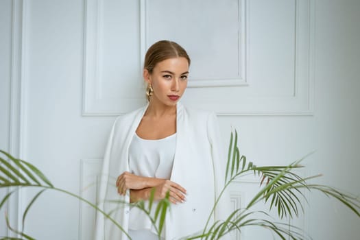 portrait of a beautiful young blonde woman with large gold earrings and a white jacket on the background of the white interior of the living room. The model in daylight and looks into the camera