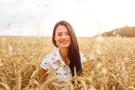 Beautiful young woman in wheat field. Cute brunette girl of Caucasian appearance in casual clothes is resting, happy in a ripe wheat field on a sunny day