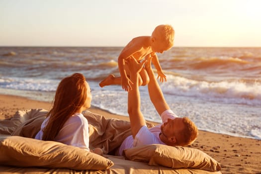 Dad tosses his son up. Lie on a bed by the sea at sunset. Family vacation on the sandy beach. Young family have fun on summer vacation.