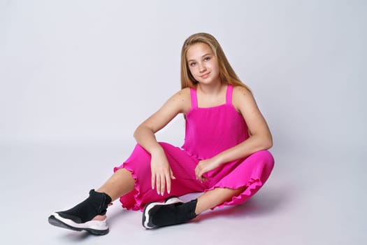 Young cute girl with long blond hair of Caucasian appearance posing in a pink suit on a white background sitting. Stylish and fashionable clothes for a teenager