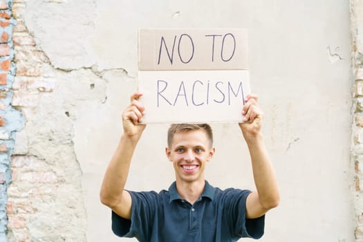 Caucasian guy came out to protest against racism with poster in his hands. Young man appearance stands against background of cement wall with cardboard in his hand on which it is written no to racism