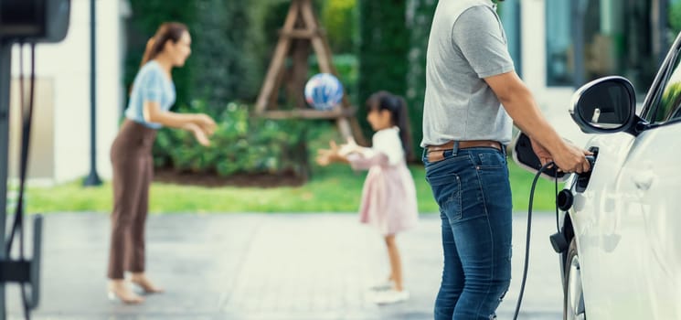 Focus image of progressive man charging electric car from home charging station with blur mother and daughter playing together in the background.