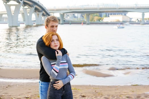 Young couple of man and woman with long red hair of Caucasian ethnicity, in casual clothes, stand on the bank of the river embracing happy on a summer day against the background of the cityscape