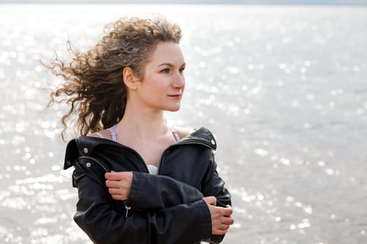 Closeup portrait of a woman, Caucasian woman with curly hair in a black leather jacket looks to the side on the shore against the background of water