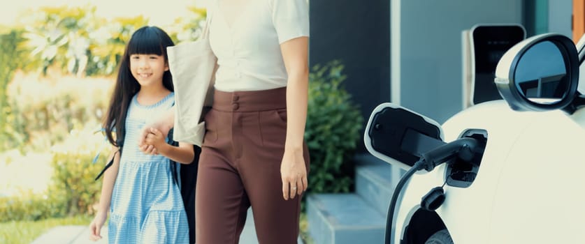 Focus electric vehicle recharging at home charging station plugged in with EV charger device with blurred background of progressive mother and daughter walking as concept for sustainability of energy.