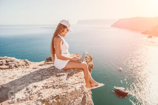 Successful business woman in yellow hat working on laptop by the sea. Pretty lady typing on computer at summer day outdoors. Freelance, travel and holidays concept.