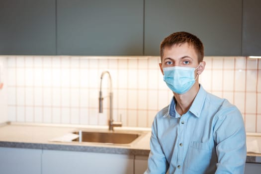 a young guy of Caucasian appearance sits in a light blue shirt and a medical mask in the kitchen alone at the table and ponders