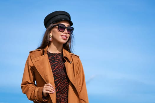 Female portrait of a beautiful young woman of Caucasian appearance in a black cap against a blue sky in sunglasses and a jacket