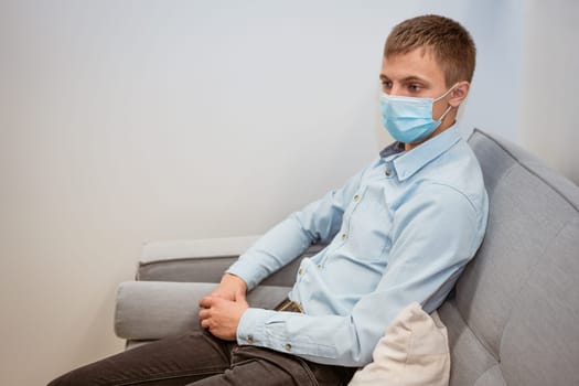 a young guy of Caucasian ethnicity in a shirt and a protective mask sits on the couch alone at home. Concept of being at home during global quarantine